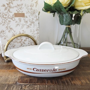 Peut inclure: Petit plat à gratin en céramique blanche avec couvercle. Le plat a une bande de couleur marron et les mots "Mini Casserole Micro" imprimés sur le côté. Le plat est posé sur une surface en bois.