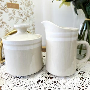 Peut inclure: Un sucrier crème avec un couvercle et une crémière assortie. Les deux pièces ont une bande blanche horizontale. Le sucrier est rond et la crémière a une anse et un bec. L'ensemble est sur un napperon en dentelle blanche.