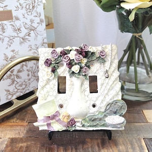 Könnte beinhalten: Ein dekorativer Lichtschalter mit einer weißen Vase mit lila und weißen Rosen. Die Abdeckung hat zwei Schalteröffnungen und ist mit einem Hut, einem Puder und einem Band verziert. Der Hintergrund hat ein Blumenmuster.