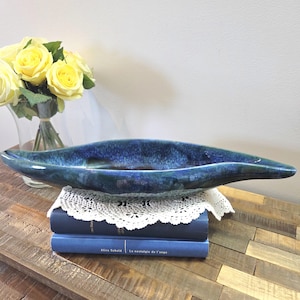 Peut inclure: Un long bol décoratif en céramique bleue avec une finition brillante. Le bol est placé sur une pile de livres bleus et un napperon en dentelle blanche. Un bouquet de roses jaunes est en arrière-plan.