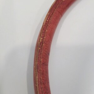 May include: Close-up of a curved, red leather handle. The handle shows signs of wear, with visible stitching and areas where the leather is worn. The background is a plain white surface.