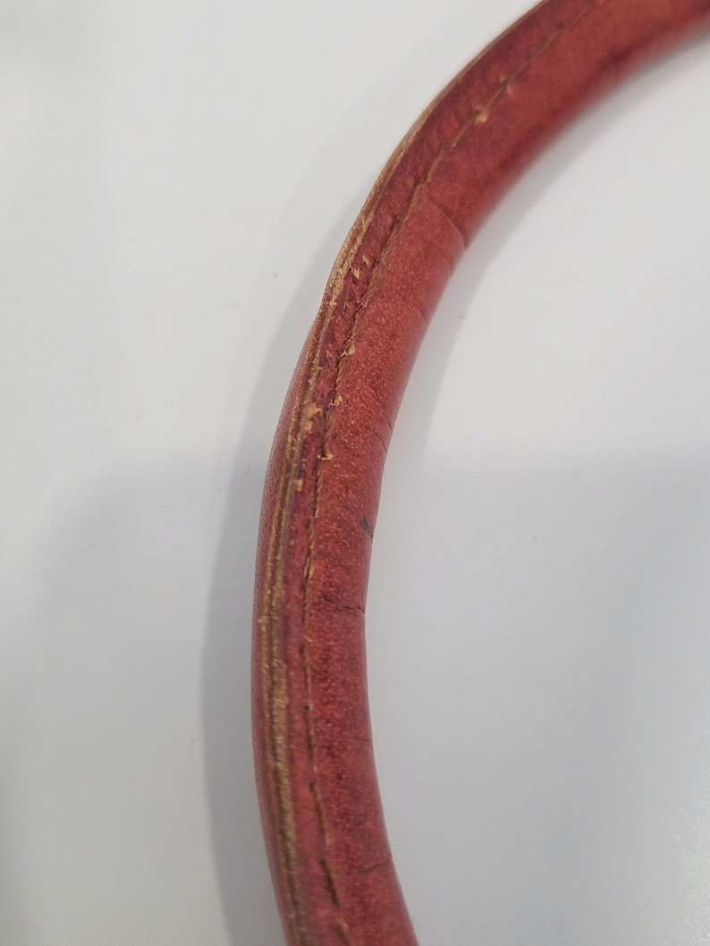 May include: Close-up of a curved, red leather handle. The handle shows signs of wear, with visible stitching and areas where the leather is worn. The background is a plain white surface.