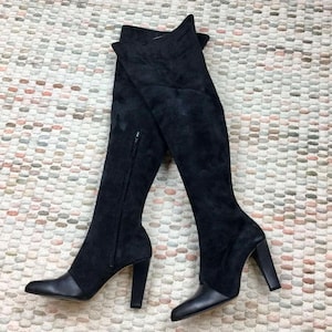 May include: Black over-the-knee boots with a suede-like texture and a pointed toe. The boots have a block heel and a zipper on the side. The boots are displayed on a patterned rug.