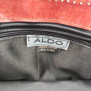May include: Close-up of the interior of a red leather handbag. The bag has a black zipper and a tag that reads "ALDO" and "GENUINE LEATHER MADE IN INDIA". The interior lining is dark gray.