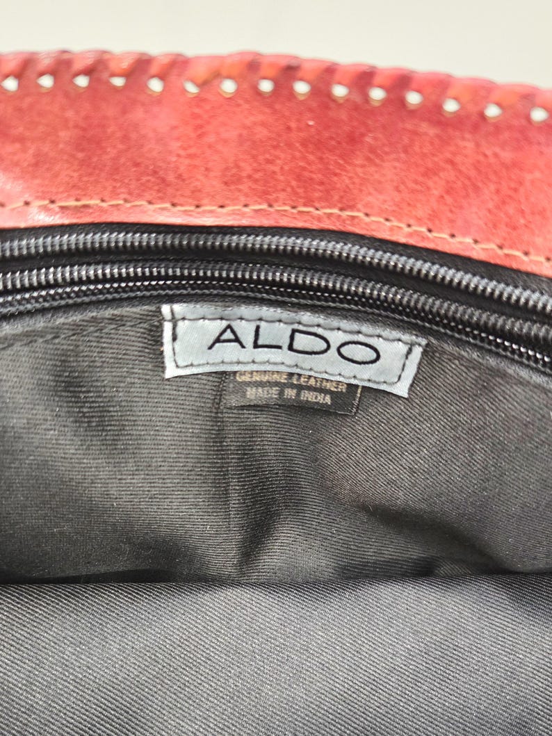 May include: Close-up of the interior of a red leather handbag. The bag has a black zipper and a tag that reads "ALDO" and "GENUINE LEATHER MADE IN INDIA". The interior lining is dark gray.