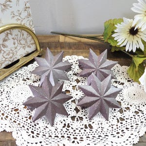Peut inclure: Quatre décorations en forme d'étoile avec une finition métallique texturée. Les étoiles sont d'une couleur gris-violet clair et reposent sur un napperon en dentelle blanche. Un arrangement floral et un cadre photo décoratif sont en arrière-plan.