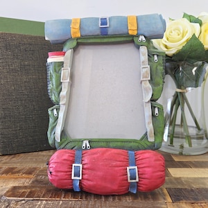 May include: A novelty photo frame designed as a green and tan backpack, complete with a blue sleeping bag and rolled mat. The frame has a rectangular aperture for a photograph and includes pockets, straps, and buckles. A red and white water bottle is visible.