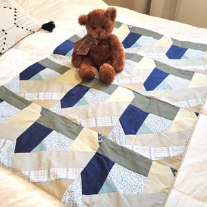 Peut inclure: Un ours en peluche marron avec une écharpe marron est assis sur une courtepointe patchwork. La courtepointe présente des motifs géométriques dans des tons de bleu, beige et gris. La courtepointe est sur un couvre-lit crème. Un oreiller décoratif avec un pompon noir est en arrière-plan.