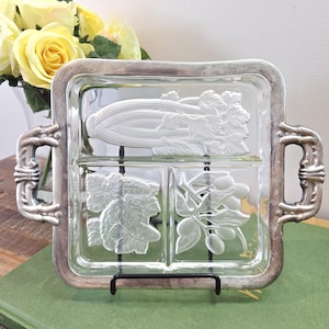 Peut inclure: Un plateau de service en verre transparent avec un cadre et des poignées en métal argenté. Le plateau est divisé en trois sections, chacune présentant des motifs en relief de légumes : céleri, feuilles et olives. Le plateau est soutenu par un support en métal noir.