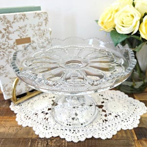 Peut inclure: Un présentoir à gâteaux en verre transparent doté d'un plateau circulaire avec un motif floral pressé, un bord festonné et une base sur pied robuste. La surface du plateau présente un motif central en étoile entouré d'alvéoles arrondies.