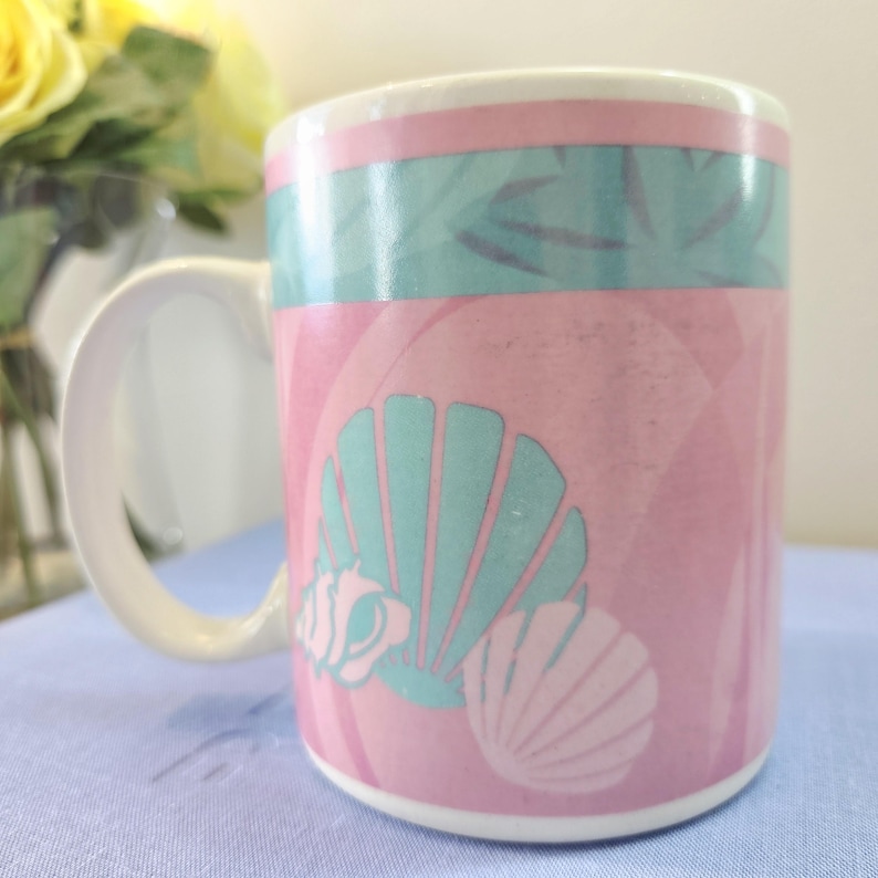 Op de afbeelding: Een keramische mok met een witte handgreep en rand. De mok heeft een roze achtergrond met turquoise schelpmotieven en een turquoise en roze band rond de bovenkant. De mok is waarschijnlijk voor dranken.