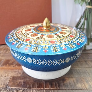 Peut inclure: Une boîte décorative vintage bleue et blanche avec un couvercle. Le couvercle présente un motif floral détaillé en rouge, or et crème. Le récipient a un bouton doré et repose sur une base blanche.