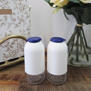 Puede incluir: Dos tarros de vidrio blanco y transparente con tapas azules. Los tarros tienen forma redondeada y están sobre una superficie de madera. En el fondo se aprecia un estampado floral decorativo.