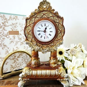 May include: An ornate, gold-toned mantel clock with a white clock face and black Roman numerals. The clock has a decorative, detailed design with a brown base and silver accents. The clock is set on a wooden surface.