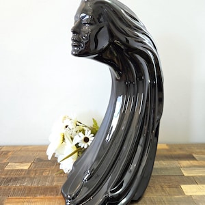 May include: A glossy black ceramic sculpture of a woman's profile with flowing hair. The sculpture has a curved, elongated form and sits on a wooden surface. White flowers are placed near the base of the sculpture.
