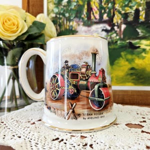 May include: A white ceramic mug with a gold rim and handle, featuring a colorful illustration of a steam roller. The mug has the text "STEAM ROLLER by AVELING 1893" at the base. The background shows a landscape scene.