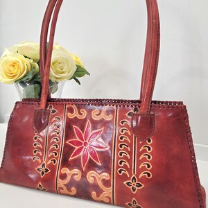May include: A reddish-brown leather handbag with two handles. The bag features a central floral design in red and tan, with decorative patterns on either side. The stitching is visible, and the bag has a rectangular shape.