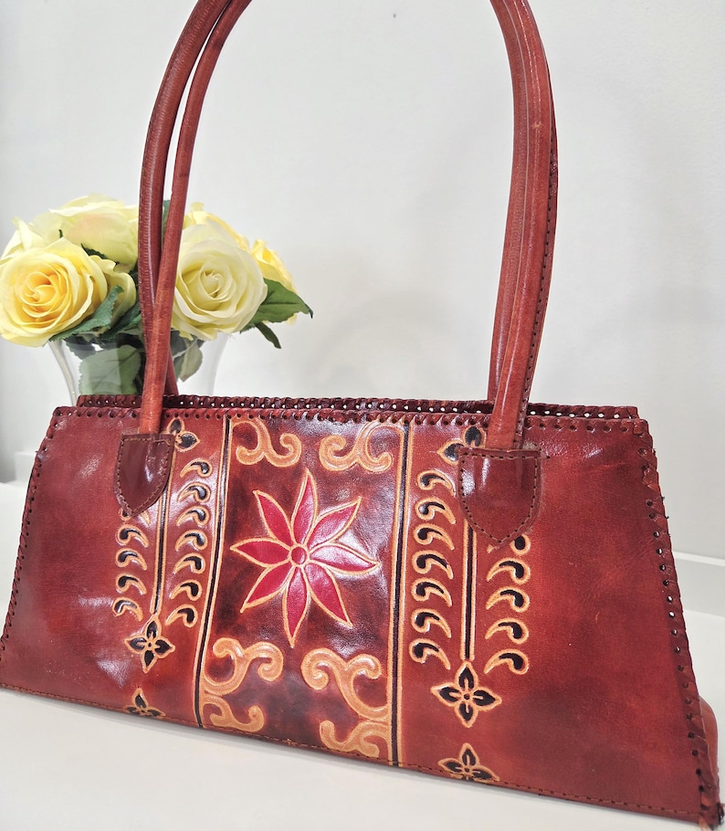 May include: A reddish-brown leather handbag with two handles. The bag features a central floral design in red and tan, with decorative patterns on either side. The stitching is visible, and the bag has a rectangular shape.