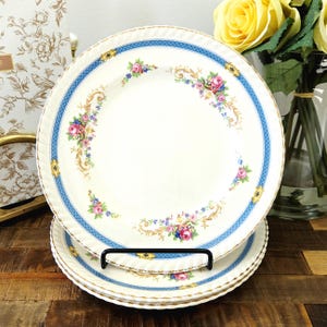 May include: A set of round ceramic plates featuring a cream-coloured centre with a floral pattern of pink roses and blue accents. The rim is decorated with a blue geometric border, gold scrollwork, and a scalloped edge with a gold-toned trim.