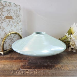 May include: A pale blue ceramic vase with a wide, shallow form. The vase has a glossy finish and is placed on a rustic wooden surface. A floral notebook and flowers are in the background.