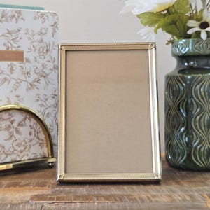 May include: A rectangular picture frame with a thin, metallic gold-toned border featuring a subtle beaded texture along the inner edge. The frame is empty, displaying a plain, light brown backing material within its vertical orientation.