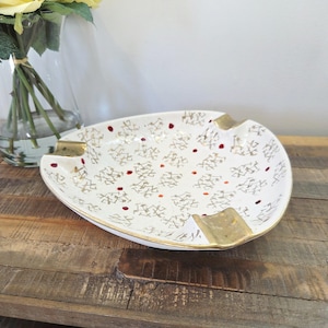May include: A vintage ashtray with a white base, gold trim, and a pattern of gold and red accents. The ashtray has a unique shape with four resting areas and is placed on a wooden surface.