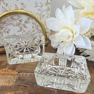 Peut inclure: Deux cendriers carrés en verre transparent avec un motif en étoile au centre et une bordure texturée. Ils reposent sur une surface en bois, avec des fleurs blanches et un élément décoratif doré en arrière-plan.