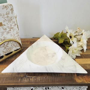 Peut inclure: Un bol décoratif triangulaire blanc avec un motif marbré. Le bol a une indentation circulaire au centre. Le bol est posé sur une surface en bois avec un arrangement floral blanc et un organisateur à motifs floraux décoratif en arrière-plan.
