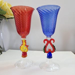Peut inclure: Deux gobelets décoratifs en verre, un rouge et un bleu. Les deux ont un motif torsadé sur le bol et des tiges ornées et colorées. Le gobelet rouge a des accents jaunes et oranges, tandis que le gobelet bleu a des accents rouges et verts.