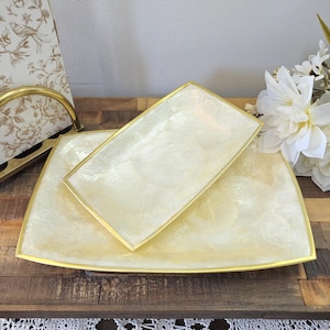Puede incluir: Dos bandejas decorativas. La bandeja más grande es rectangular con esquinas redondeadas, y la más pequeña es rectangular. Ambas bandejas tienen un acabado nacarado con ribete dorado. Las bandejas se exhiben sobre una superficie de madera.