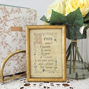 May include: A framed print with gold bamboo-style trim features French text. The text is in a decorative font with a large initial letter. A floral patterned book and a gold-toned arched object are in the background. Yellow roses are in a glass vase.