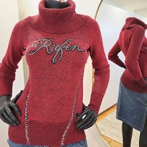 Puede incluir: Un suéter burdeos con cuello alto. La palabra "Rufen" está bordada en cursiva en el pecho. Una cadena recorre la parte delantera del suéter. La prenda se exhibe en un maniquí.