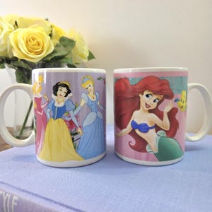 Op de afbeelding: Twee witte keramische mokken met Disney-prinsessenontwerpen. E&eacute;n mok toont Sneeuwwitje, Assepoester en Aurora. De andere mok toont Ariel uit De Kleine Zeemeermin. Beide mokken hebben witte handvatten en staan tegen een lichtpaarse achtergrond.