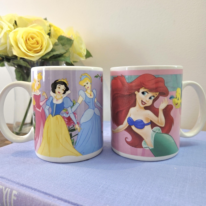 Op de afbeelding: Twee witte keramische mokken met Disney-prinsessenontwerpen. E&eacute;n mok toont Sneeuwwitje, Assepoester en Aurora. De andere mok toont Ariel uit De Kleine Zeemeermin. Beide mokken hebben witte handvatten en staan tegen een lichtpaarse achtergrond.