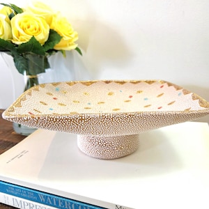 Peut inclure: Plat à servir rectangulaire en céramique crème, avec un socle. Le plat présente un motif texturé et pointillé avec des bordures dorées et des accents colorés. Une pièce décorative pour servir des aliments ou exposer des objets.