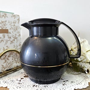 Peut inclure: Une cafetière noire vintage avec une bande dorée au milieu. La cafetière a un corps arrondi, une poignée noire et un couvercle noir. Elle repose sur un napperon en dentelle blanche. Un carnet floral et des fleurs sont en arrière-plan.