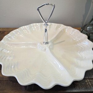 Puede incluir: Una fuente para servir dividida de color crema con borde festoneado. La fuente tiene tres secciones y un asa plateada. El asa tiene forma triangular. La fuente para servir está sobre una superficie de madera oscura.