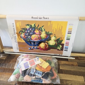 May include: A Royal Paris needlepoint canvas featuring a still life of fruit in a blue bowl, mounted on a wooden frame. The canvas includes a colour key and the text 131 83. A plastic bag containing assorted embroidery threads and needles is included.