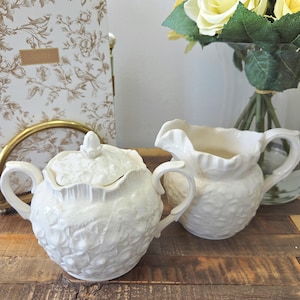 Peut inclure: Sucrier et pot à crème en céramique crème. Le sucrier a un couvercle et deux poignées. Le pot à crème a un bord festonné et une seule poignée. Un carnet et des fleurs sont en arrière-plan.