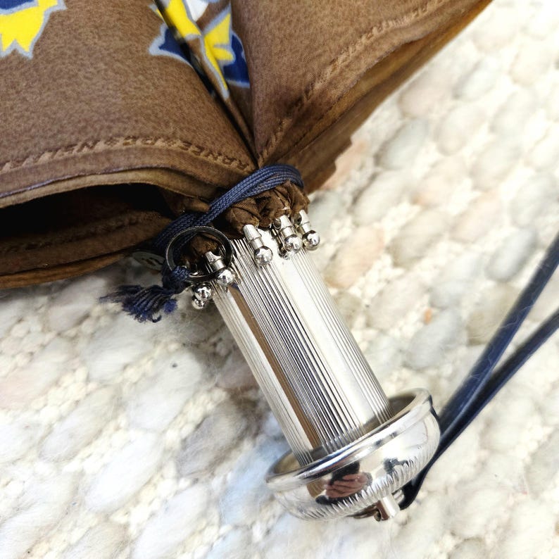 Pu&ograve; includere: Primo piano di un ombrello marrone con manico argentato. Il tessuto dell'ombrello presenta un motivo floreale giallo e blu. Il manico &egrave; scanalato e ha una base e una parte superiore decorative in argento. Un cordino blu assicura l'ombrello.