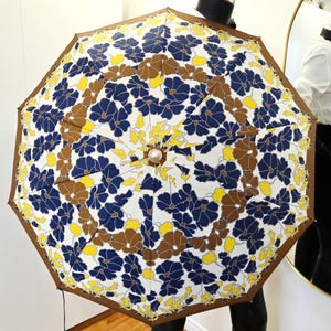 Pu&ograve; includere: Un ombrello aperto con un motivo floreale. L'ombrello presenta uno sfondo bianco con un motivo ripetuto di fiori blu, marroni e gialli. I bordi sono rifiniti con un bordo marrone.