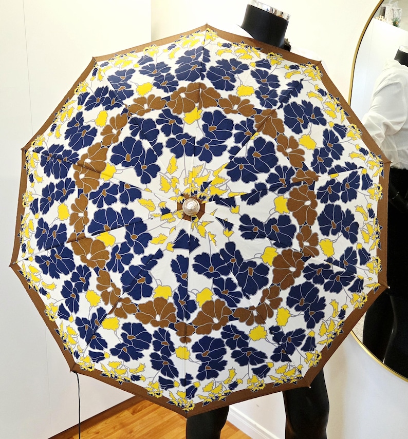 Pu&ograve; includere: Un ombrello aperto con un motivo floreale. L'ombrello presenta uno sfondo bianco con un motivo ripetuto di fiori blu, marroni e gialli. I bordi sono rifiniti con un bordo marrone.