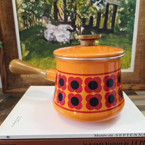 Può includere: Casseruola in smalto arancione con manico e coperchio in legno. La casseruola presenta un motivo floreale rosso e nero sulla base. Un oggetto vintage per la cucina.
