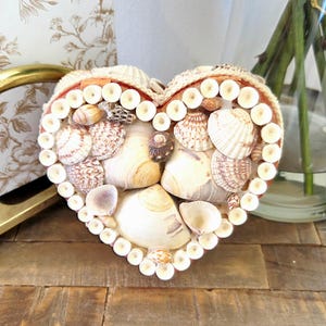 May include: A heart-shaped box decorated with a variety of seashells. The box is filled with shells in shades of white, beige, and brown. The edges are lined with small, round, white shells, creating a textured border.