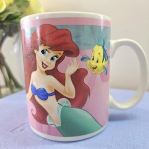 Op de afbeelding: Witte keramische mok met Ariel uit De Kleine Zeemeermin. De mok heeft een roze achtergrond met Ariel, een blauwogige zeemeermin met rood haar en een gele vis. De mok heeft een wit handvat en is geschikt voor warme of koude dranken.