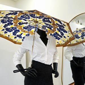 Pu&ograve; includere: Un ombrello aperto con un motivo floreale blu navy, giallo e marrone. &Egrave; tenuto da un manichino nero che indossa una camicia bianca e una gonna nera. Il motivo dell'ombrello si riflette in uno specchio.