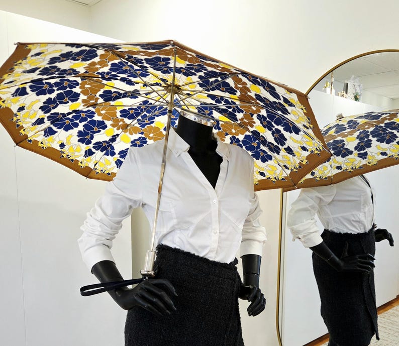 Pu&ograve; includere: Un ombrello aperto con un motivo floreale blu navy, giallo e marrone. &Egrave; tenuto da un manichino nero che indossa una camicia bianca e una gonna nera. Il motivo dell'ombrello si riflette in uno specchio.