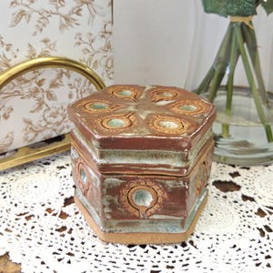 May include: A hexagonal ceramic lidded box with a glossy brown and light blue glaze. The lid and sides feature embossed circular floral patterns. The base of the container is unglazed, showing the natural tan clay color of the pottery piece.