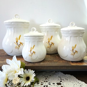 May include: Set of four white ceramic canisters with lids. Each canister features a wheat stalk design in shades of yellow and brown. The canisters are arranged on a wooden surface, with a white floral arrangement and lace doily in the foreground.