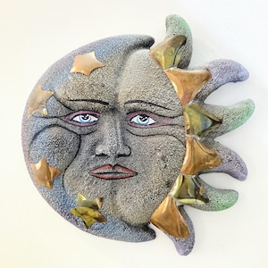 Peut inclure: Décoration murale céleste représentant un visage de croissant de lune avec des yeux bleus, des lèvres rouges et des étoiles en cuivre. Les rayons du soleil sont un mélange de cuivre, de vert et de violet. L'œuvre a un aspect texturé en pierre.